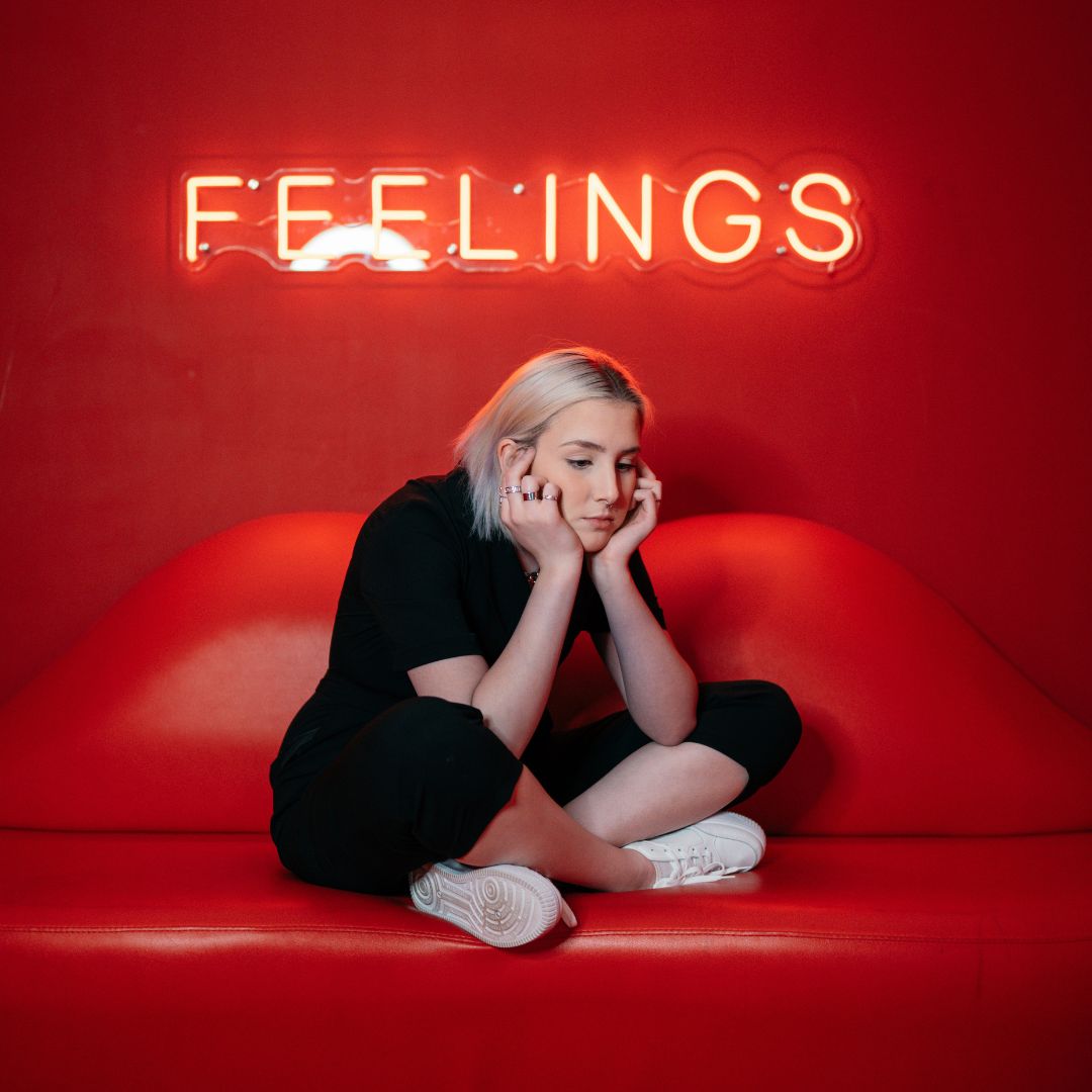 dziewczyna blondynka siedzi zamyślona po turecku na czerwonej kanapie w kształcie ust na ścianie za nią napis feelings