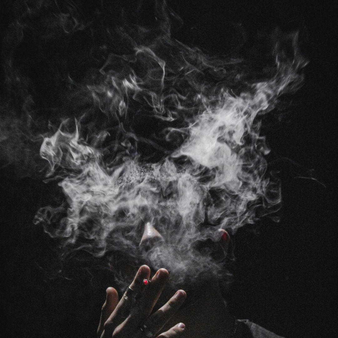 mężczyzna w czarnej bluzce na czarnym tle z zapalonym papierosem w dłoni z twarzą ukrytą za papierosowym dymem